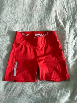 Röda träningsshorts från Swix - Snygga röda träningsshorts från Swix med bred resår i midjan och vit logga framtill. Shortsen har en sportig passform och är tillverkade i ett stretchigt syntetmaterial som passar perfekt för aktiva dagar.