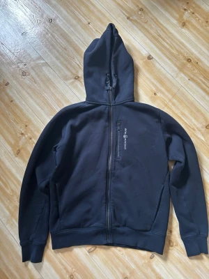 Svart hoodie från Sail Racing  - Svart hoodie med dragkedja från Sail Racing. Klassisk modell med huva och två fickor framtill. Diskret logga på bröstet och mjukt material som känns skönt mot huden. Perfekt för en avslappnad och sportig stil.