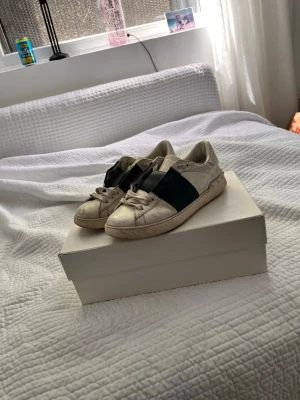 Valentino open - Snygga vita sneakers från Valentino Garavani med bred svart läderdetalj över mitten. Klassisk låg modell med rund tå och vita skosnören. Tillverkade i läder för en exklusiv känsla och stilren look.