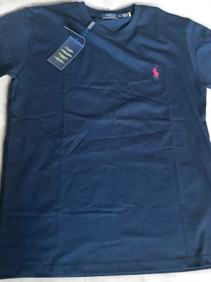 Svart t-shirt från Polo Ralph Lauren - Snygg svart t-shirt från Polo Ralph Lauren med klassisk röd logga broderad på bröstet. T-shirten är kortärmad och har en rund halsringning. Tillverkad i mjuk bomull för en skön känsla.