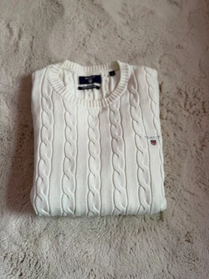 Vit kabelstickad tröja från Gant - Vit kabelstickad tröja från Gant med rund halsringning och broderad logotyp på bröstet. Tröjan har långa ärmar och ribbade muddar vid ärmslut och nederkant. Klassisk och stilren design i mjukt material.
