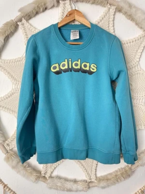 Adidas tröja  - Cool turkos sweatshirt från Adidas med rund halsringning och tryckt logga i gult och svart framtill. Tröjan har långa ärmar och är gjord i mjukt bomullsmaterial. Perfekt för en avslappnad och sportig stil.