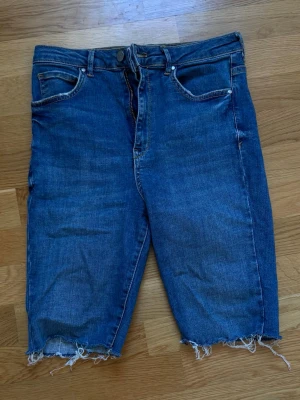 Blå jeansshorts från Gina Tricot - Säljer ett par klassiska blå jeansshorts från Gina Tricot med råa, fransiga benslut och femficksmodell. Shortsen har normal passform och stängs med dragkedja och knapp. Perfekta för sommaren och lätta att matcha med allt. Klippt själv