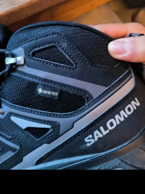 Svarta Salomon Gore-Tex kängor - Säljer ett par svarta kängor från Salomon med Gore-Tex-material som håller fötterna torra. Snygg design med grå detaljer och Salomon-logga på sidan. Perfekta för dig som gillar outdoor och vill ha något slitstarkt och bekvämt.