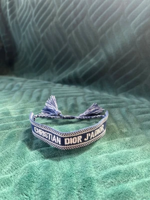 Blått broderat armband Dior - Snyggt blått armband från Christian Dior med texten 'CHRISTIAN DIOR J'ADIOR' broderad i vitt. Armbandet är gjort i textil och har tofsar i ändarna för justerbar passform. Perfekt accessoar för att lyfta din outfit.