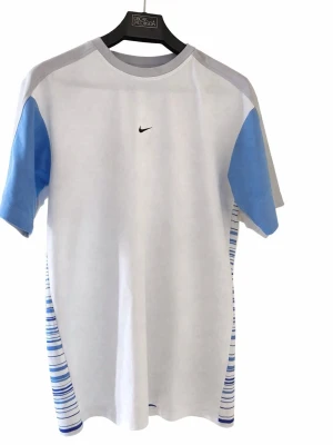 Nike vit t-shirt med blå detaljer - Nike t-shirt i vitt med ljusblå ärmar och grå axelparti. Clean Nike t-shirt träningsmaterial.