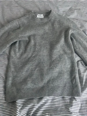 Grå stickad tröja från j lindberg  - Klassisk grå stickad tröja från j Lindberg med rund halsringning och ribbade muddar vid ärmslut och nederkant. Tröjan har en enkel och stilren design, perfekt för lager på lager och passar till det mesta. Materialet är mjukt och bekvämt.