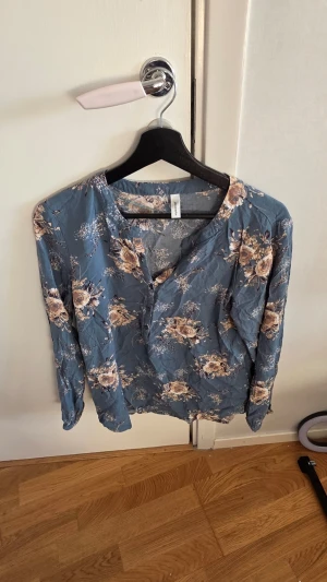 Blommig blå blus från Soyaconcept - Supersöt blus i blått med beige och vita blommor från Soyaconcept. Blusen har rund halsringning, knappar framtill och långa ärmar. Tunt och lätt material som känns luftigt och skönt. Perfekt för dig som gillar romantiska och feminina plagg.