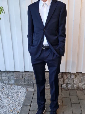 Marinblå kostym från Dressmann - Snygg mörkblå kostym från Dressmann i slim fit-modell. Kavajen har klassisk passform med två knappar och byxorna är raka. Kostymen är iprincip oanvänd och har inga defekter. Kostymen är köpt för 2500 kr. Kostymöverdelen är i storlek 44 short och kostymbyxorna är i storlek 31-32. På bilden är jag cirka 175 cm. Priset kan diskuteras, hör av dig vid minsta fråga!