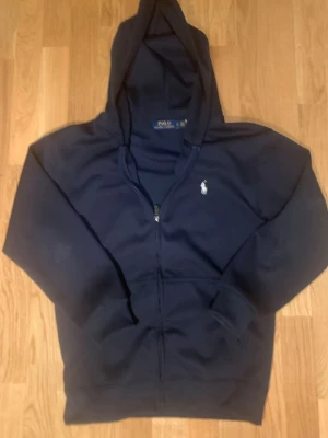 Marinblå hoodie från Polo Ralph Lauren - Snygg marinblå hoodie från Polo Ralph Lauren med dragkedja framtill och klassisk vit logga på bröstet. Hoodien har huva och fickor på sidorna. Tillverkad i mjukt bomullsmaterial som är skönt att bära.