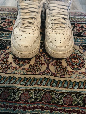 Nike Air Force 1 vita sneakers - Klassiska vita Nike Air Force 1 sneakers med svart swoosh på sidan. Skorna har lågt skaft, perforerad tåbox och är tillverkade i läder med gummisula. Perfekta för dig som gillar streetwear och tidlös design.