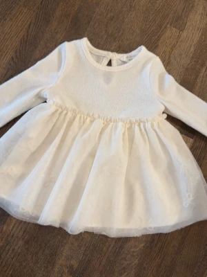 Vit klänning med tyll och rosetter - Supersöt vit klänning från Primark med lång ärm och fluffig kjol i tyll. Kjolen har små broderade rosetter och prickar för en extra gullig look. Perfekt för speciella tillfällen eller när du vill känna dig extra fin. Size 0-3months