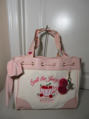 Juicy Couture daydreamer tote rosa och vit  - Söt handväska från Juicy Couture i rosa och vitt med broderad text 'Spill the Juicy' och körsbärsmotiv. Väskan har rosa detaljer, guldiga metalldelar, en stor rosett på sidan och en fluffig nyckelring. Materialet är en mix av tyg och konstläder.