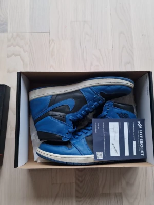 Nike Air Jordan 1 Retro High OG blå - Säljer ett par Nike Air Jordan 1 Retro High OG sneakers i blått och svart läder med vit sula. Klassisk high-top siluett med ikoniska Swoosh-loggan på sidan och Air Jordan Wings-märket vid ankeln. Perfekta för dig som gillar streetwear och sneakerskultur.