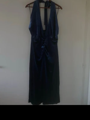 Mörkblå långklänning i satin - Snygg mörkblå långklänning i glansigt satinmaterial med djup v-ringning och knyt i nacken. Klänningen har en elegant omlottkänsla framtill och är ärmlös med öppen rygg. Perfekt för dig som vill sticka ut på nästa event.