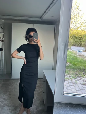 Svart långklänning med slits - Stilren svart långklänning med korta ärmar och slits framtill. Klänningen har en figurnära passform och är perfekt för dig som gillar en enkel men elegant look. Tillverkad i ett mjukt och stretchigt material som sitter snyggt på kroppen.