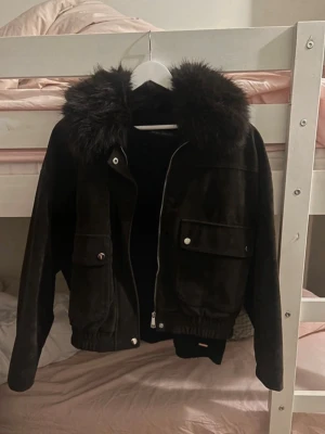 Mocka pälsjacka - Snygg svart skinnjacka med stor krage i fuskpäls och dragkedja framtill. Jackan har två stora fickor med lock och silverfärgade knappar samt resår i midjan. Perfekt för dig som vill ha en trendig och varm look.