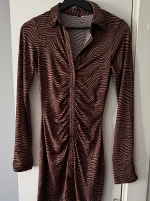 Brun mönstrad långärmad klänning från H&M - Snygg brun klänning från H&M med vågigt mönster och knappar framtill. Klänningen har lång ärm, krage och rynkade detaljer längs knappraden. Materialet känns stretchigt och mjukt, perfekt för dig som gillar en trendig look.