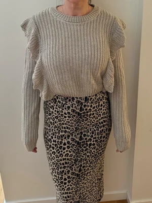 Beige stickad tröja med volanger - Trendig beige stickad tröja med volangdetaljer längs axlar och sidor. Grovstickad struktur och rund halsringning. Perfekt för dig som vill ha en unik och snygg stickad tröja till garderoben.