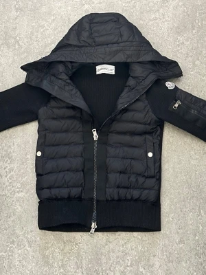 Svart dunjacka med huva från Moncler - Snygg svart dunjacka från Moncler med stickade ärmar och rygg, quiltad front och stor huva. Jackan har dragkedja framtill, två fickor med tryckknappar och en liten ficka med dragkedja på ärmen. Perfekt för kalla vinterdagar.