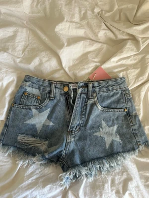 Jeansshorts med stjärnor från Design by Amor - Trendiga ljusblå jeansshorts från Design by Amor med slitna detaljer, fransig kant och stora stjärnor på framsidan. Klassisk femficksmodell och hög midja. Perfekta för sommardagar och festivaler.