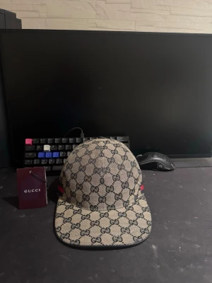 Gucci beige keps med GG-mönster - Säljer en mörkblå/beige keps från Gucci med det klassiska GG-monogrammönstret över hela kepsen. Kepsen har en böjd skärm och detaljer i rött och blått på sidan. Märkets logga syns tydligt och materialet känns lyxigt och slitstarkt.