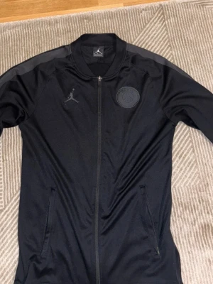 Svart PSG x Jordan ziptröja - Svart ziptröja från Jordan i samarbete med PSG. Tröjan har diskreta loggor på bröstet, ribbad krage och dragkedja framtill. Snygga detaljer på axlarna och två fickor med dragkedja. Perfekt för en sportig och stilren look.