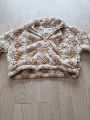 Beige och vit teddytröja från Hollister - Supermjuk stickad teddytröja från Hollister i beige och vitt rutmönster. Tröjan har halv dragkedja framtill, hög krage och avslappnad passform. Perfekt för kalla dagar när du vill vara både varm och snygg.