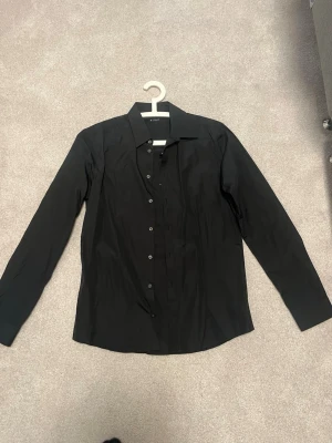 Svart skjorta från Eton, storlek 41 - Klassisk svart skjorta från Eton med lång ärm och knappar framtill. Skjortan har en stilren design med krage och är tillverkad i ett slätt bomullstyg. Perfekt för dig som gillar en clean och tidlös look.