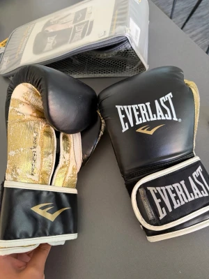 Everlast boxningshandskar - Säljer ett par Everlast Powerlock boxningshandskar i svart och guld. Handskarna har bred kardborreknäppning och är tillverkade i syntetmaterial. Perfekta för träning och sparring, med cool design och bra passform.