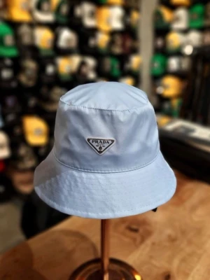 Ljusblå bucket hat från Prada - Snygg ljusblå bucket hat från Prada med klassisk siluett och ikonisk logotyp framtill. Hatten är tillverkad i ett lätt och bekvämt material som ger en cool och avslappnad vibe. Perfekt accessoar för att lyfta din streetstyle.