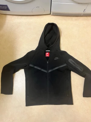 Svart Nike hoodie med dragkedja - Barn storlek btw Svart hoodie från Nike med hel dragkedja framtill och huva. Snygga detaljer med svarta ränder över bröstet och en praktisk ficka med dragkedja på ena ärmen. Perfekt för en sportig och avslappnad stil.