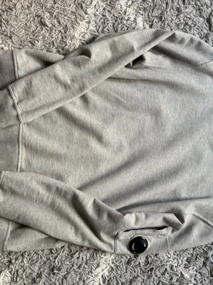 Grå sweatshirt från C.P. Company - Snygg grå sweatshirt från C.P. Company med klassisk passform och en unik detalj – en svart lins på ena ärmen. Tillverkad i mjukt bomullsmaterial som känns skönt mot huden. Perfekt för dig som gillar stilrena och moderna plagg.