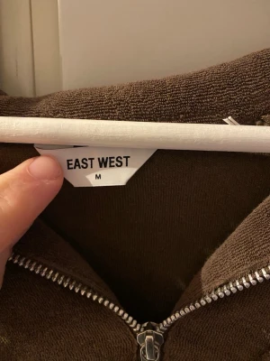 Tjocktröja  från East west  - Brun hoodie från East West i storlek M. Tillverkad i mjukt tyg med dragkedja framtill och ficka på bröstet. Perfekt för en avslappnad och stilren look. Klassisk modell med huva och ribbade muddar. 