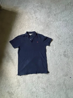 Mörkblå pikétröja från Abercrombie - Klassisk mörkblå pikétröja från Abercrombie & Fitch med korta ärmar, krage och knappar framtill. Liten röd logga broderad på bröstet. Perfekt till jeans eller shorts för en avslappnad look.61 kr frakt bitalas av köpare.