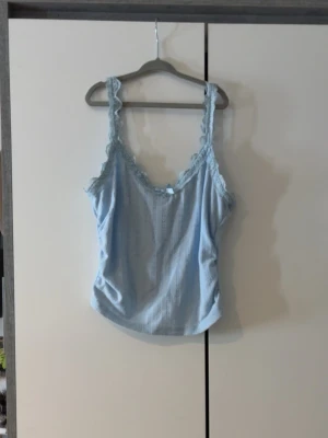 Ljusblå spetskantad linnetopp - Supersöt ljusblå linnetopp med spetskantade axelband och en liten rosett framtill. Toppen har en feminin v-ringning och är tillverkad i ett mjukt, lätt material som passar perfekt till sommaren.