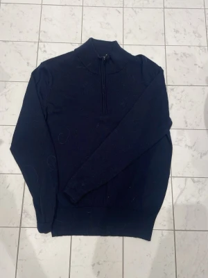 Mörkblå halvzip stickad tröja Uniqlo - Snygg mörkblå stickad tröja från Uniqlo med halvzip och hög krage. Tröjan har långa ärmar, ribbade muddar och är tillverkad i ett mjukt, tunt material som passar perfekt till lager på lager. Enkel och stilren design.