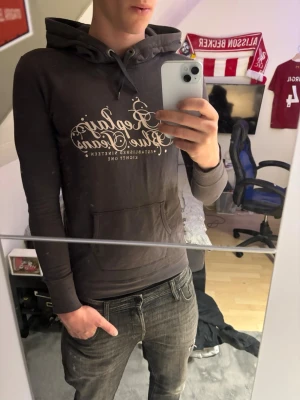Mörkgrå Replay Blue Jeans hoodie - Snygg mörkgrå hoodie från Replay med stor vit text 'Replay Blue Jeans' på bröstet och klassisk känguruficka. Tröjan har huva med dragsko och är tillverkad i mjukt bomullsmaterial. Perfekt för en avslappnad och trendig stil. Storlek M men sitter som S 