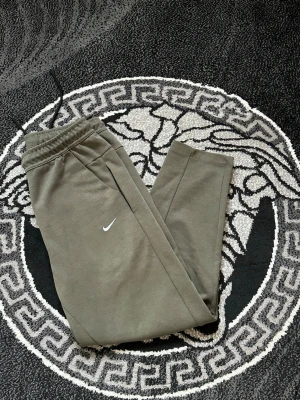 Olivgröna Nike mjukisbyxor - Snygga olivgröna mjukisbyxor från Nike med klassisk vit swoosh på låret. Byxorna har elastisk midja med svart dragsko och praktiska sidofickor. Perfekt för chill eller träning, tillverkade i mjukt material som känns skönt mot huden.
