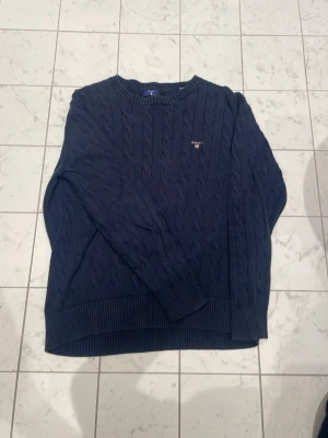 Mörkblå kabelstickad tröja GANT - Snygg mörkblå kabelstickad tröja från GANT med rund halsringning och broderad logga på bröstet. Tröjan har ribbade muddar vid ärmslut och nederkant, perfekt för kalla dagar. Klassisk och tidlös design som passar till det mesta. En för 200kr båda för 300kr