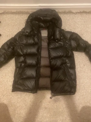 Svart dunjacka från Moncler - (Äkta) haft den i två år har knappt använt den Snygg svart dunjacka från Moncler med glansig finish och vadderad design. Jackan har huva, två stora dragkedjefickor framtill och Moncler-logga på insidan. Perfekt för kalla vinterdagar och riktigt trendig look.