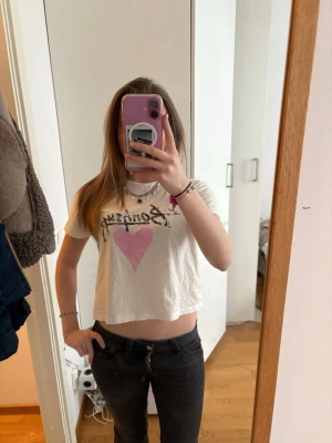 Vit croppad t-shirt med tryck - Säljer en vit croppad t-shirt med svart texten 'Bonjour' och ett stort rosa hjärta på bröstet. T-shirten har korta ärmar och en avslappnad passform. Perfekt för dig som gillar en söt och trendig stil.