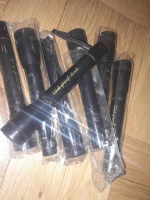 Svarta ficklampor Waterproof Torch - Säljer flera svarta ficklampor med texten 'Waterproof Torch' i guld. De är tillverkade i metall och har en smal, avlång form. Ficklamporna har av/på-vred och vissa är fortfarande i plastförpackning. Perfekt att ha i väskan eller bilen.