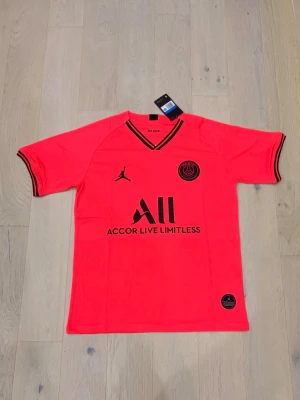 Neonrosa PSG matchtröja Jordan - Snygg neonrosa PSG matchtröja från Jordan med svarta detaljer och loggor. Tröjan har korta ärmar, V-ringad hals med svart kant och är tillverkad i ett lätt och ventilerande material, perfekt för träning eller match.