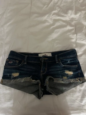 Mörkblå low waist jeansshorts från Hollister - Säljer ett par mörkblå jeansshorts från Hollister med slitna detaljer och råa kanter. Klassisk femficksmodell med låg midja och snygga kontrastsömmar. Perfekta för sommaren. Storleken är w25, midjemåttet är ca 35-37cm