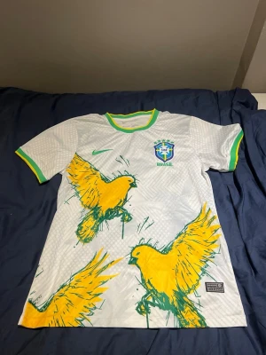 Vit Brasilien matchtröja Nike - Snygg vit Brasilien matchtröja från Nike med gröna och gula detaljer på krage och ärmslut. Framsidan har stora gula fåglar och Brasilien-emblemet på bröstet. Tillverkad i lätt och ventilerande material, perfekt för fotboll eller träning.