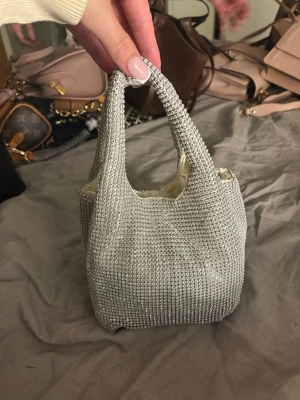 Silvrig kuvertväska med pärlor - Säljer en silvrig kuvertväska täckt av små glittrande pärlor. Väskan har en mjuk, fyrkantig form och två handtag. Insidan är fodrad i ljus textil. Perfekt för dig som vill ha en unik accessoar med extra bling.