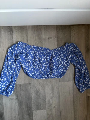 Blå off shoulder topp med blommor - Trendig blå off shoulder topp med vita småblommiga mönster och långa puffärmar. Toppen har resår vid axlar och midja för en snygg passform. Perfekt för dig som gillar en romantisk och somrig stil. Använd ett fåtal gånger.