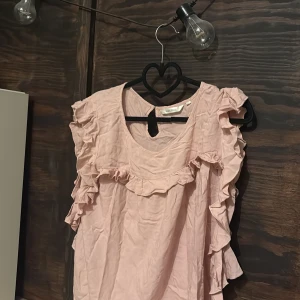 Ljusrosa volangtopp från Ally Bell - Supersöt ljusrosa topp från Ally Bell med volangdetaljer längs ärmar och framstycke. Toppen har rund halsringning och är ärmlös, perfekt för en trendig och feminin look. Tillverkad i mjukt bomullsmaterial. 