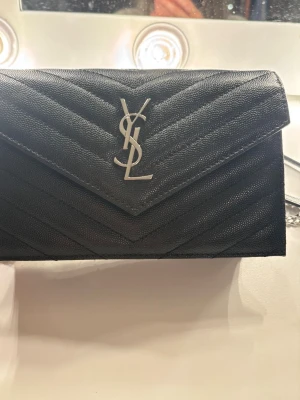 Svart YSL handväska med kedjerem - Säljer en svart handväska från Saint Laurent med klassiskt YSL-logo i silver på framsidan. Väskan har ett kuvertformat lock, quiltat mönster och en snygg silverfärgad kedjerem med läderdetalj. Tillverkad i läder och perfekt för dig som gillar stilrena accessoarer.